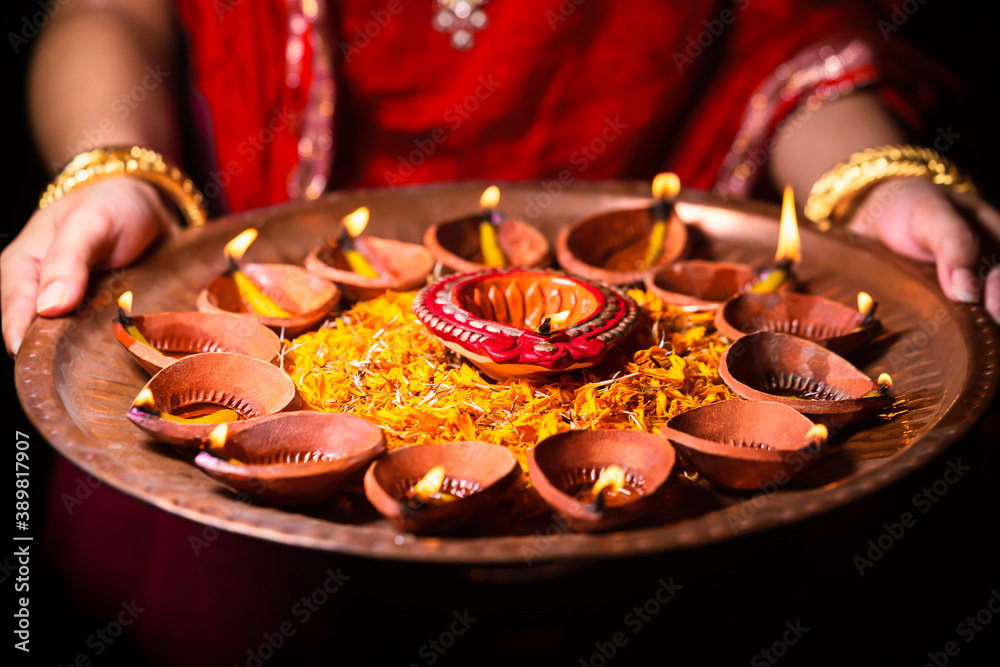 Woman Holding Puja Thali. Happy Diwali Background. Indian woman or bride wearing traditional red cloth and jewelry, holding lit burning diya or clay oil lamps, decorated with flowers.
