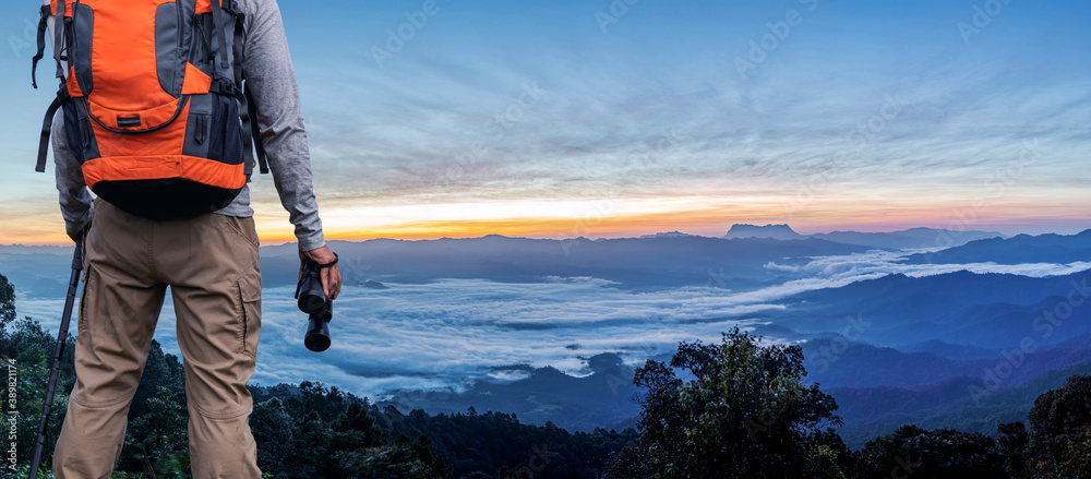 Young man with backpack and holding a binoculars standing on mountain looking foggy