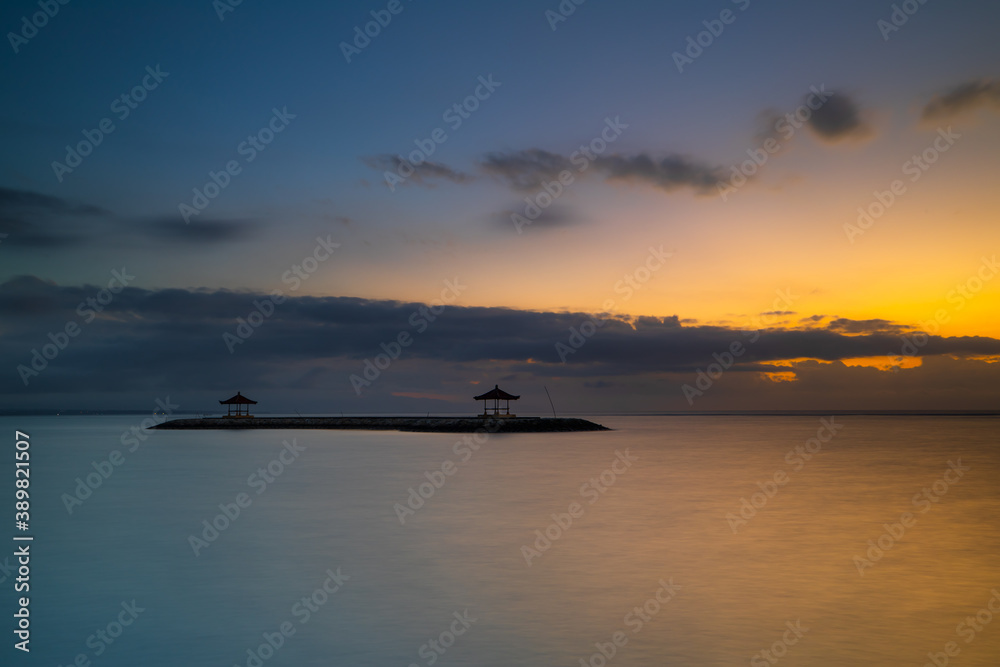 Sunrise view. Seascape. Mountains and Agung volcano. Traditional gazebos on an artificial island in the ocean. Water reflection. Sanur beach, Bali, Indonesia.