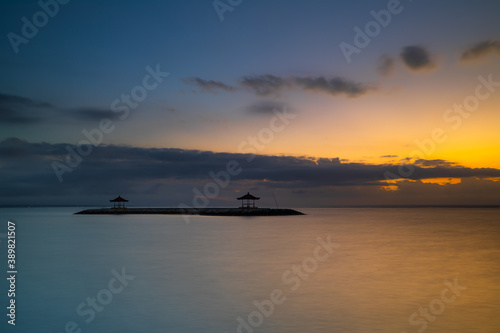 Sunrise view. Seascape. Mountains and Agung volcano. Traditional gazebos on an artificial island in the ocean. Water reflection. Sanur beach, Bali, Indonesia.