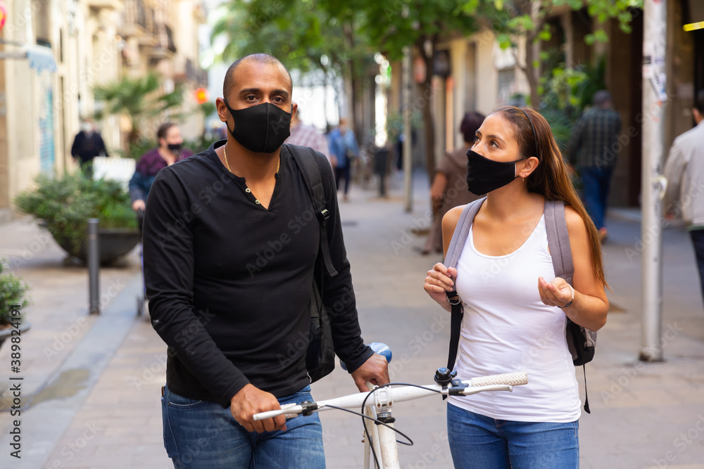 Young hispanic couple wearing black protective masks to prevent viral infection walking with bike along city street on warm fall day