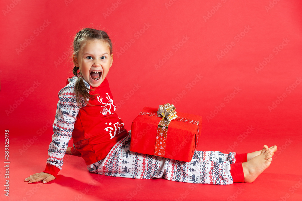 laughing girl with two pigtails and in Christmas pajamas sits on the floor and holds a gift box on her lap, isolated on a vibrance background.