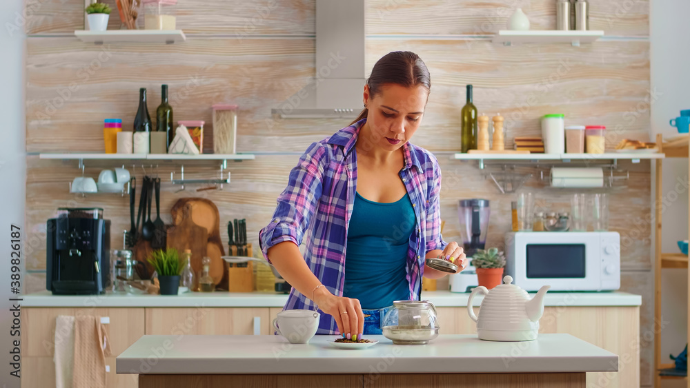Fototapeta premium Young woman preparing green tea for breakfast in a modern kitchen using teapot sitting near the table. Putting with hands, pressing herbal, healthy, tea leaves, in pot, in the morning.