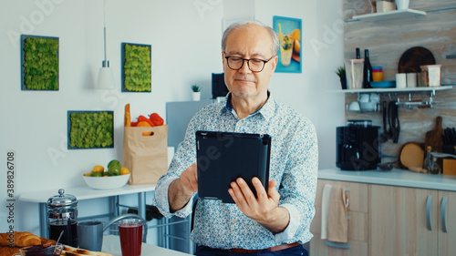 Happy older man in kitchen during breakfat using tablet pc. Elderly person with tablet portable pad PC in retirement age using mobile apps, modern internet online information technology with photo