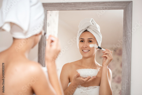 Woman applying facial clay mask. Beauty treatments. Close-up portrait of beautiful girl with a towel on her head applying facial mask, Spa procedures at home