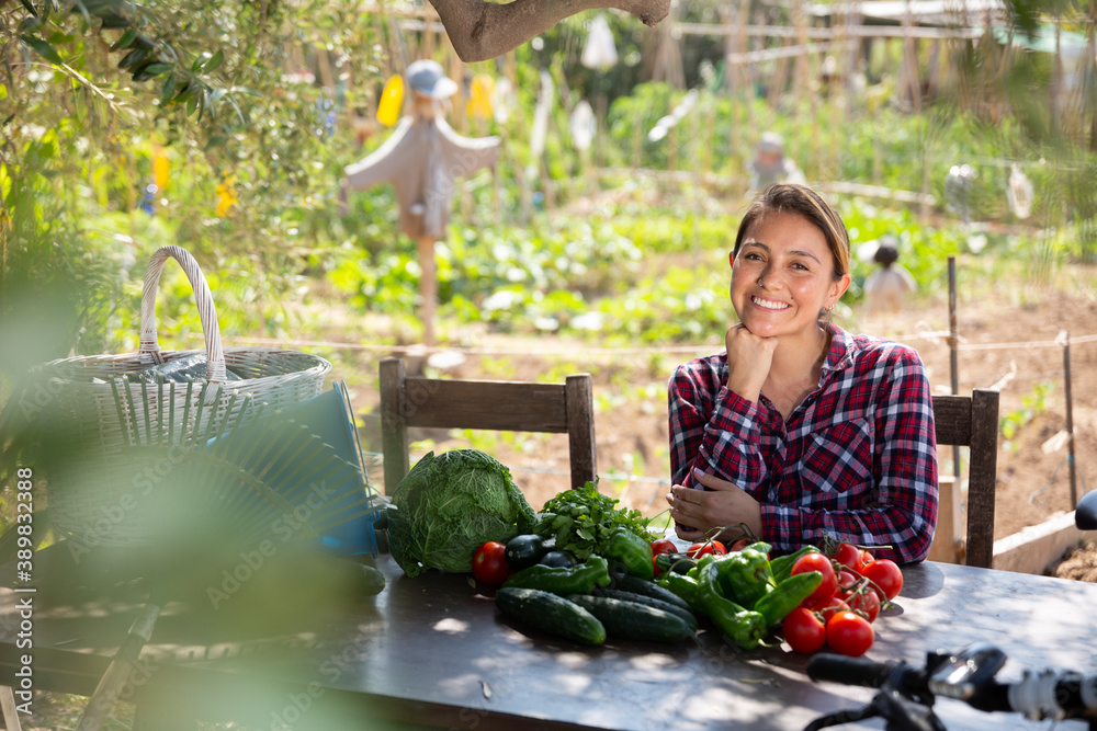Positive young woman sits at table with ripe vegetables in the backyard of village house