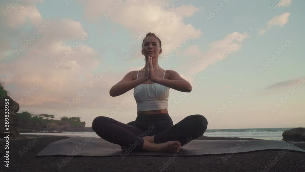 Full low angle arc of sun-tanned Caucasian girls wearing sports attire sitting in lotus pose joining palms in namaste on massive stone by ocean shore. Female meditator practicing at sunset Full low angle arc of sun-tanned Caucasian girls wearing sports attire sitting in lotus pose joining palms in namaste on massive stone by ocean shore. Female meditator practicing at sunset