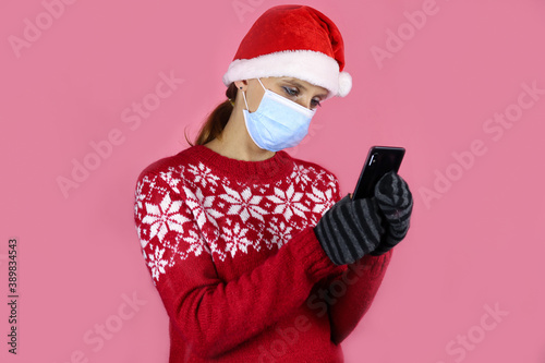 A young girl in santa claus hat and medical protective mask looks into a mobile phone