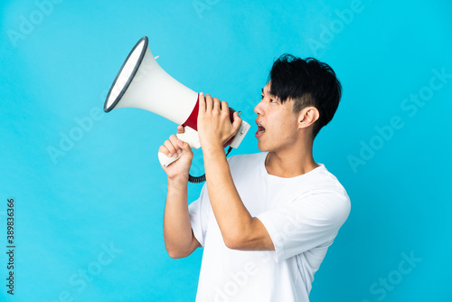 Young Chinese man isolated on blue background shouting through a megaphone to announce something in lateral position photo