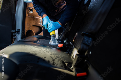 The interior of the car is being cleaned. A worker uses a special vacuum cleaner to clean the black car seat. The worker is wearing blue rubber gloves.