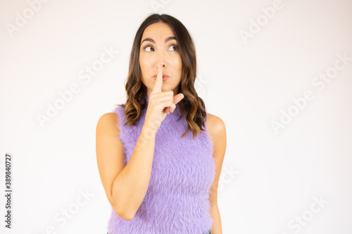 Portrait of beautiful and young brunette woman standing over isolated white background asking to be quiet with finger on lips. Silence and secret concept.