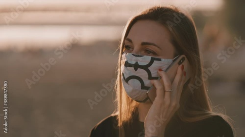 Beautiful young woman with a mask on is having conversation on the phone. She is standing on the beach during amizing sunset. photo