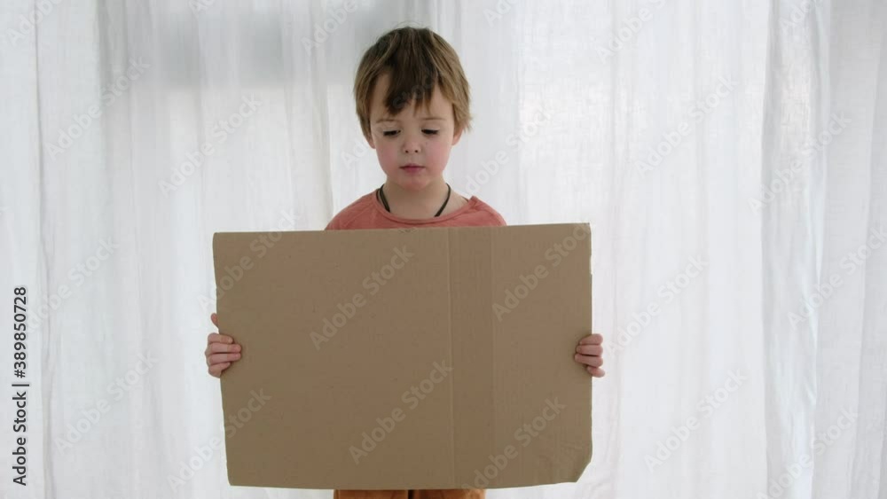 Upset little boy in orange holds large cardboard sheet with space for template standing near white curtain in light room Upset little boy in orange holds large cardboard sheet with space for template standing near white curtain in light room