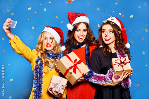 Three smiling women in santa helper hats with many gift boxes make selfie. Young women posing on blue background. Christmas, new year, winter, party concept.
