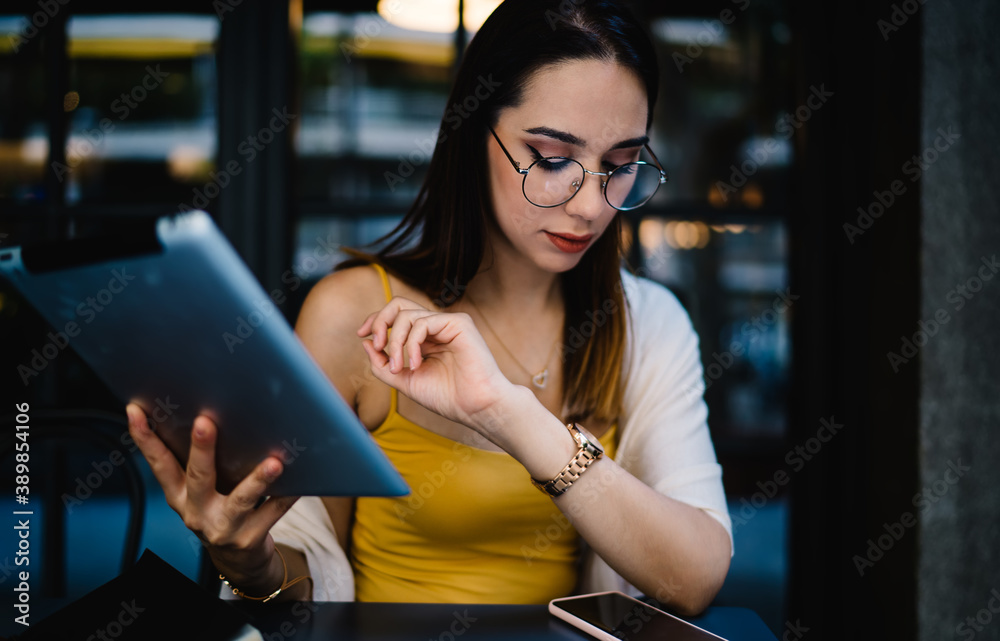 Serious young female freelancer checking time while working with tablet in outdoor cafe