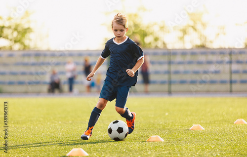 Young boy in blue soccer jersey uniform running after ball on training pitch....