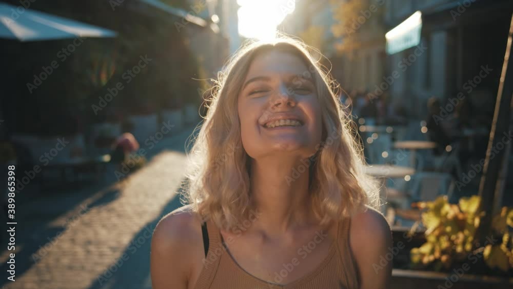 Enjoying every moment! Close up portrait of funny caucasian blonde travel women smiling into camera – having fun and laughing on the street of old city with paving stone. Sun behind her head Enjoying every moment! Close up portrait of funny caucasian blonde travel women smiling into camera – having fun and laughing on the street of old city with paving stone. Sun behind her head