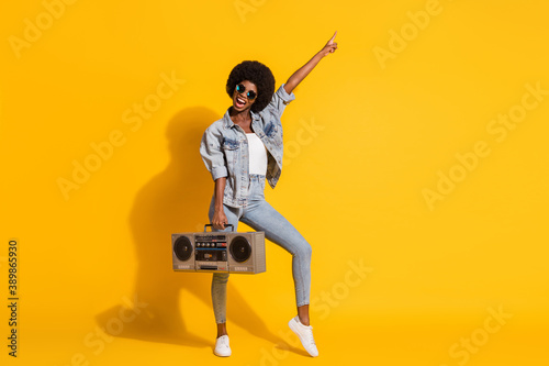 Full length photo of adorable lady wear casual denim outfit dancing boombox pointing empty space isolated yellow color background