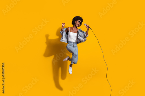 Full length photo of adorable young lady wear casual denim outfit jumping singing microphone isolated yellow color background