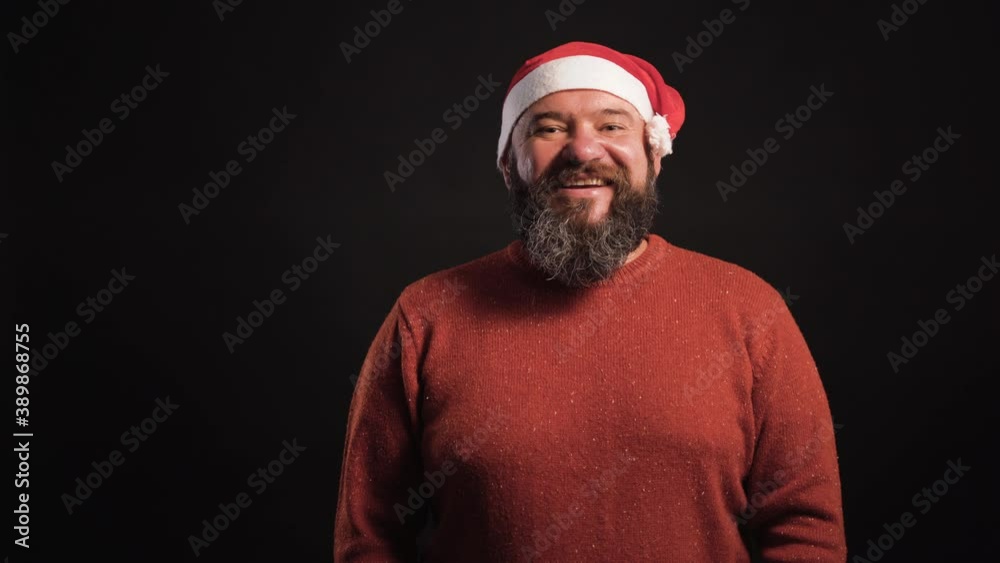 A bearded man on a black background in a santa claus hat lifts a box rewound with a red ribbon A bearded man on a black background in a santa claus hat lifts a box rewound with a red ribbon