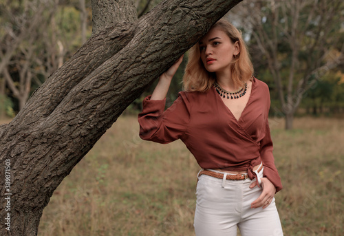 Beautiful blonde woman in in a brown blouse and white trousers posing in the forest