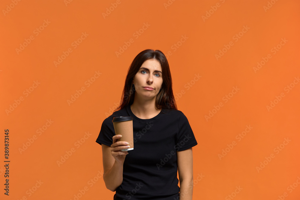 I asked for coffee and this is tea. Pensive displeased caucasian woman in black t-shirt, looks sadly at camera, holds takeaway coffee, unhappy with her drink. Isolated over an orange background. I asked for coffee and this is tea. Pensive displeased caucasian woman in black t-shirt, looks sadly at camera, holds takeaway coffee, unhappy with her drink. Isolated over an orange background.