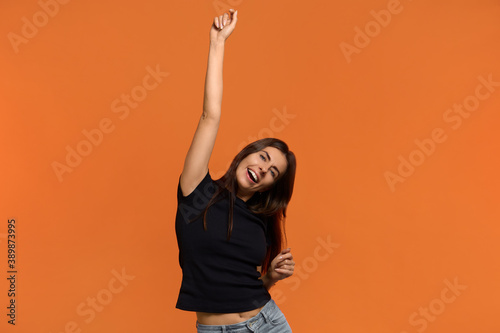 Wonderful sound. Joyful caucasian woman in black t-shirt does lucky dance, raises hands up in hooray, feels like champion after getting triumph, has fun. Isolated over an orange background. © 5M