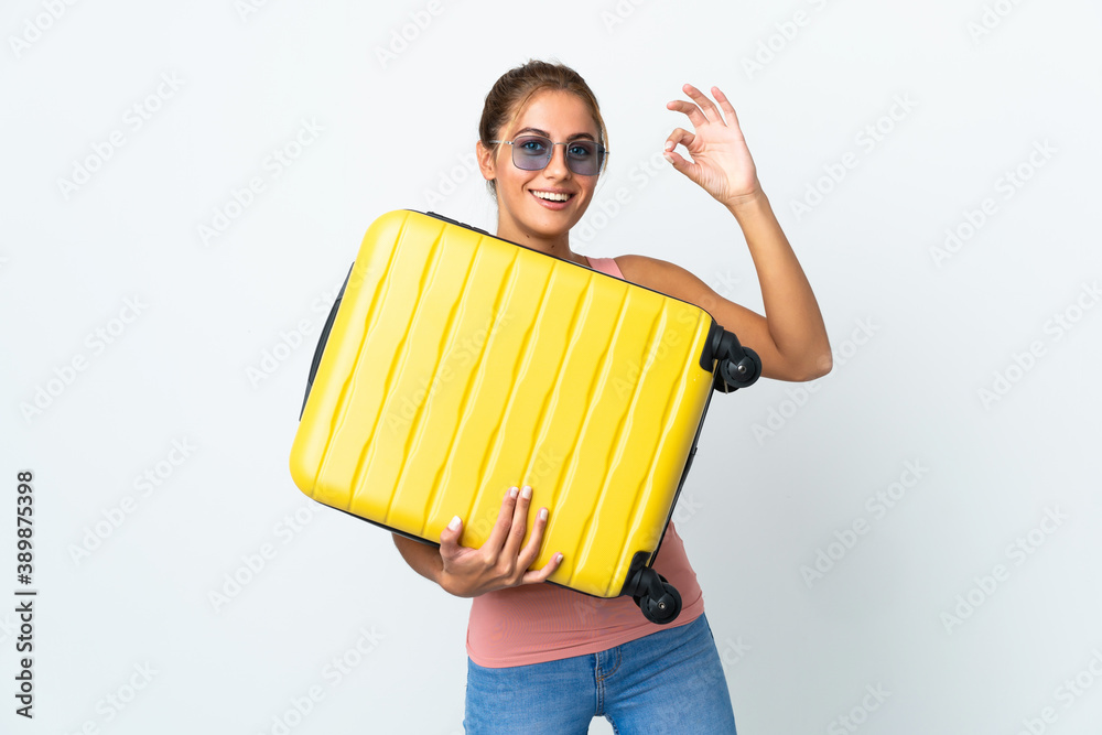 Obraz premium Young student woman isolated on white background in vacation with travel suitcase and making OK sign