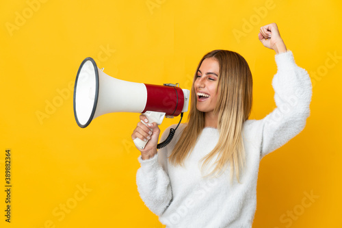 Young blonde woman isolated on yellow background shouting through a megaphone to announce something in lateral position