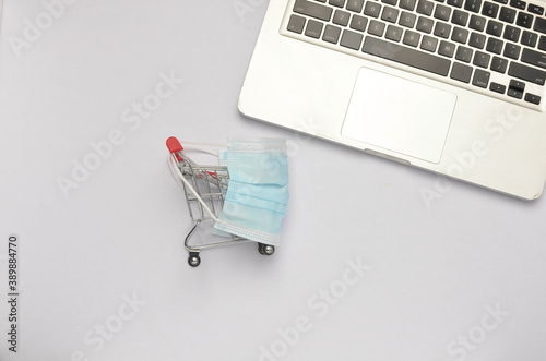 Shopping trolley and medical face mask with laptop on white background. Covid-19 pandemic. Online shopping. Selective focus.