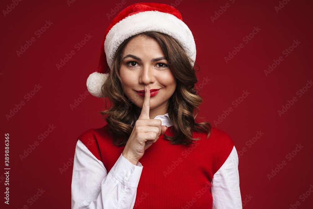 Positive young woman in christmas santa hat showing silence gesture Positive young woman in christmas santa hat showing silence gesture