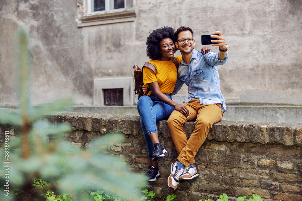 Young multiracial couple sitting on a wall and taking selfie. Young multiracial couple sitting on a wall and taking selfie.