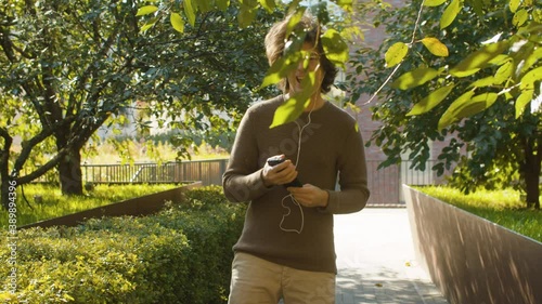 Funny hipster dancing in a park, holding smartphone in hands and listening to music through headphones. Autumn sunny day. Steadicam shot