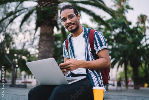 Portrait of positive dark skinned male it developer enjoying working outdoors using 4G connection and laptop computer, millennial african american student browse information for online education