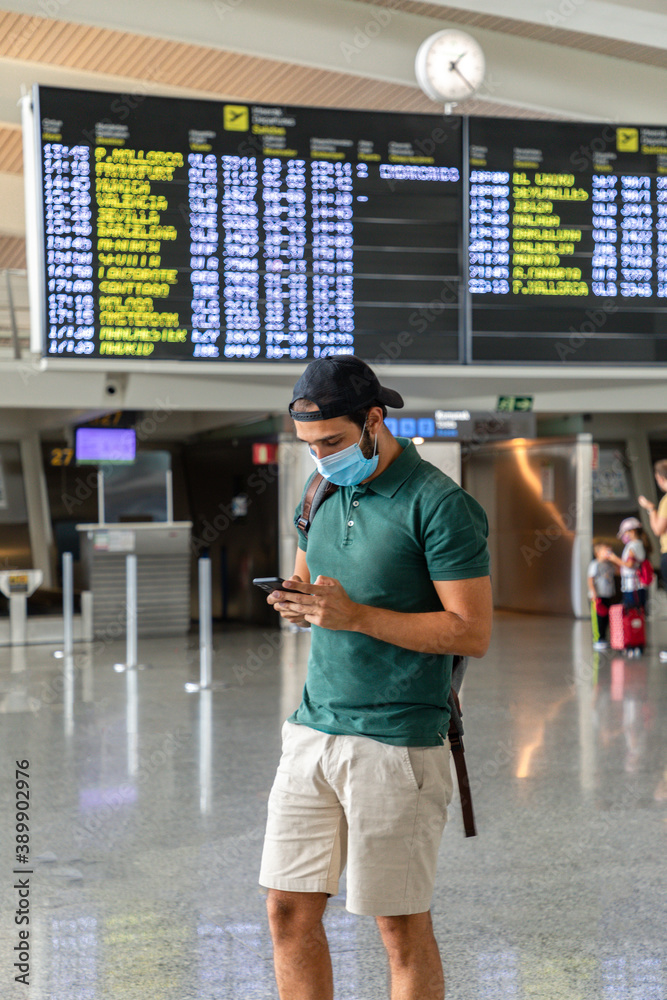 young man with mask in the airport terminal while looking at his mobile phone