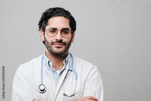 Skilled professional and adult physician dressed in white labcoat poses with crossed arms in white background.