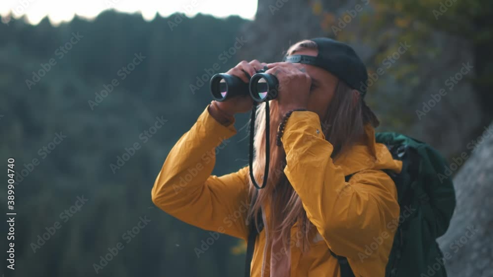 So amazing forest! Bearded tourist man with long hair in yellow jacket wearing backpack looking through binoculars while standing at the top of the hill and inspired by beautiful view in mountain So amazing forest! Bearded tourist man with long hair in yellow jacket wearing backpack looking through binoculars while standing at the top of the hill and inspired by beautiful view in mountain