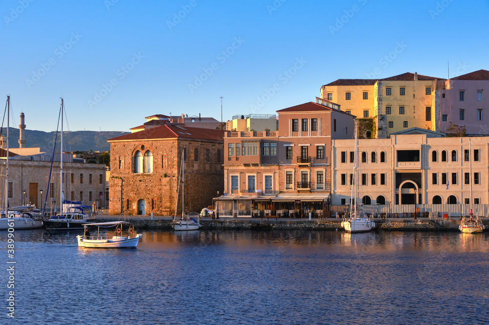 Fishing boat pass by Grand Arsenal in Old Venetian harbour, Chania, Crete, Greece. Sunrise view of quay and sailing boats anchored by piers, cafes.