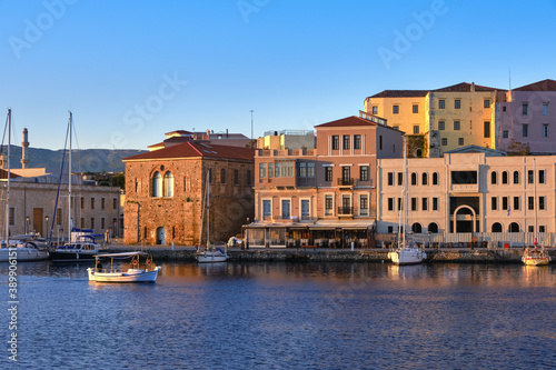 Fishing boat pass by Grand Arsenal in Old Venetian harbour, Chania, Crete, Greece. Sunrise view of quay and sailing boats anchored by piers, cafes.
