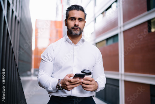 Half length portrait of confident proud CEO looking at camera during coffee break in city, Caucasian middle aged corporate director with cellular device and takeaway cup posing at urban setting