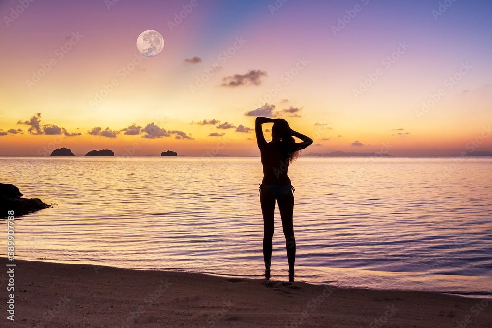 Silhouette of a young woman standing on the sandy beach by the sea against the backdrop of a spectacular sunset and a huge rising full moon on a summer evening
