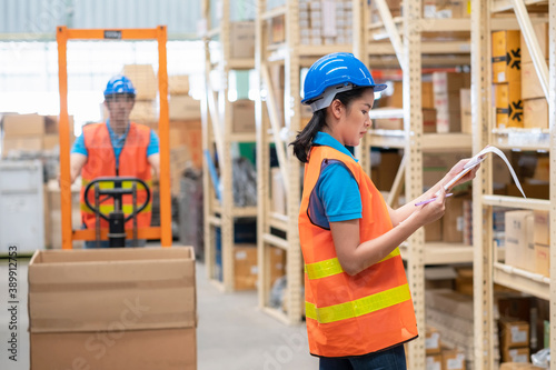 Woman worker holds clip charts and takes notes in the automotive parts warehouse distribution. Engineers wear a helmet standing in factory. In background shelves with goods and man control hand lift
