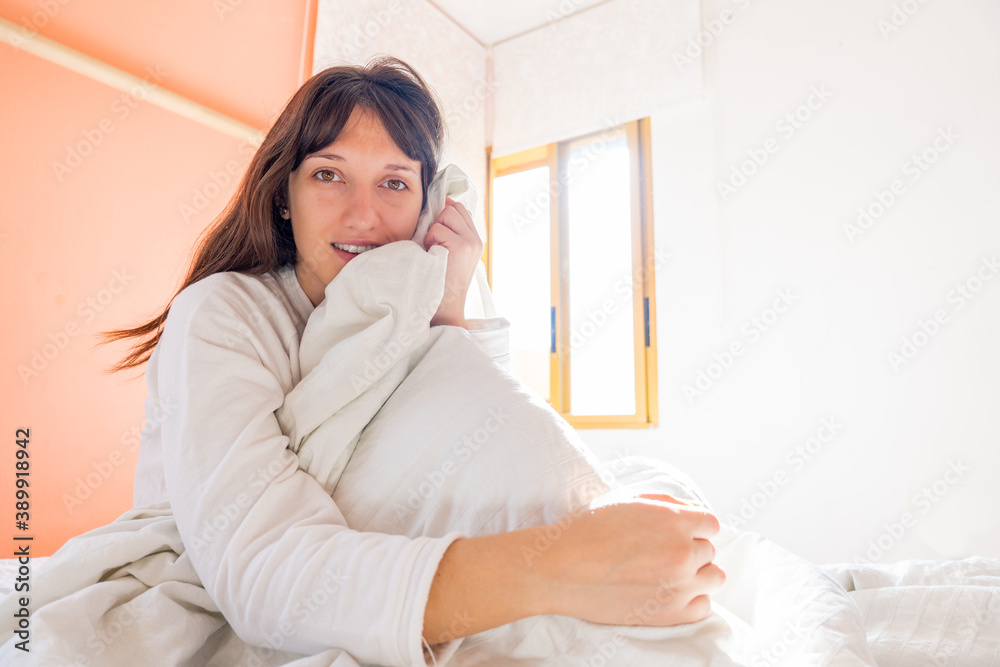 Portrait of young woman sitting on the bed of white sheets with light coming through the window behind her, pretty girl with honey colored eyes Portrait of young woman sitting on the bed of white sheets with light coming through the window behind her, pretty girl with honey colored eyes