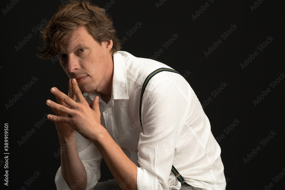 Serious thoughtful man sits in thinking position. Shaggy Caucasian guy wearing a white shirt and suspenders isolated on black background with copy space at right. High quality photo Serious thoughtful man sits in thinking position. Shaggy Caucasian guy wearing a white shirt and suspenders isolated on black background with copy space at right. High quality photo
