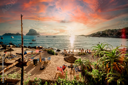 Picturesque view of Cala d'Hort tropical Beach, people hangout in beautiful beach with Es Vedra rock view during magnificent vibrant sunset glowing sun. Balearic Islands. Ibiza, Spain