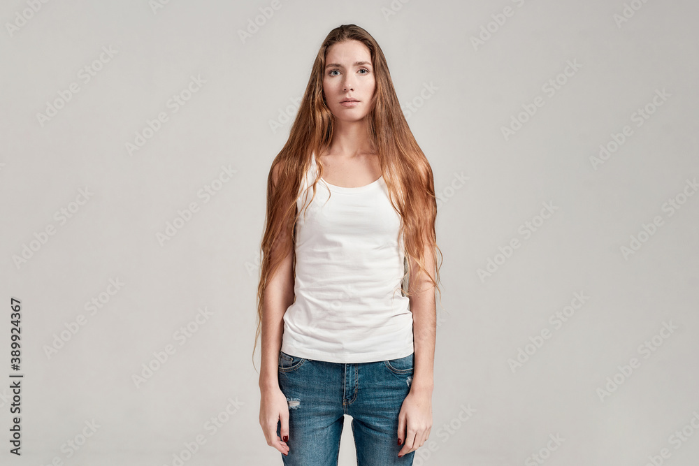 Portrait of young caucasian woman with long hair wearing white shirt and jeans looking at camera while standing isolated over grey background Portrait of young caucasian woman with long hair wearing white shirt and jeans looking at camera while standing isolated over grey background