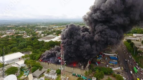 Aerial view overlooking a warehouse on fire, dark smoke rising from house ruins, sunny day, in Puerto Rico, USA - High angle, tracking, drone shot photo