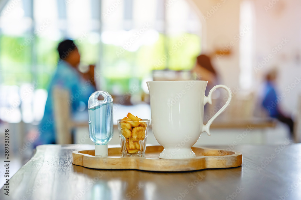 Mini portable alcohol gel bottle to kill Corona Virus(Covid-19) and biscuits and coffee cup on table with blurred image of a family eating at restaurant in background. Health care concept.