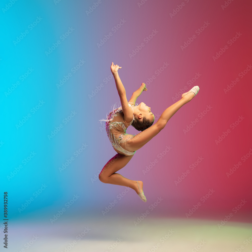 Fototapeta premium High jump. Little caucasian girl, rhytmic gymnast training, performing isolated on gradient blue-red studio background in neon. Graceful and flexible, strong child. Concept of sport, motion, action.