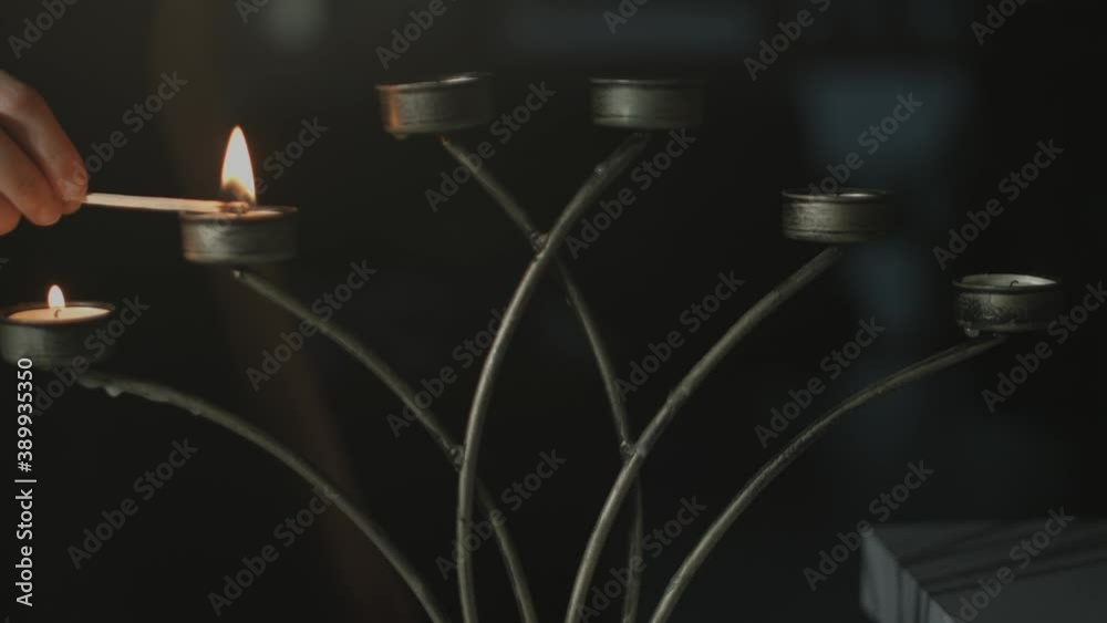 Tealights in a silver candle holder being ignited by a lit match stick by a a young woman with a black dress by night with a dark background. Shot in 4K.